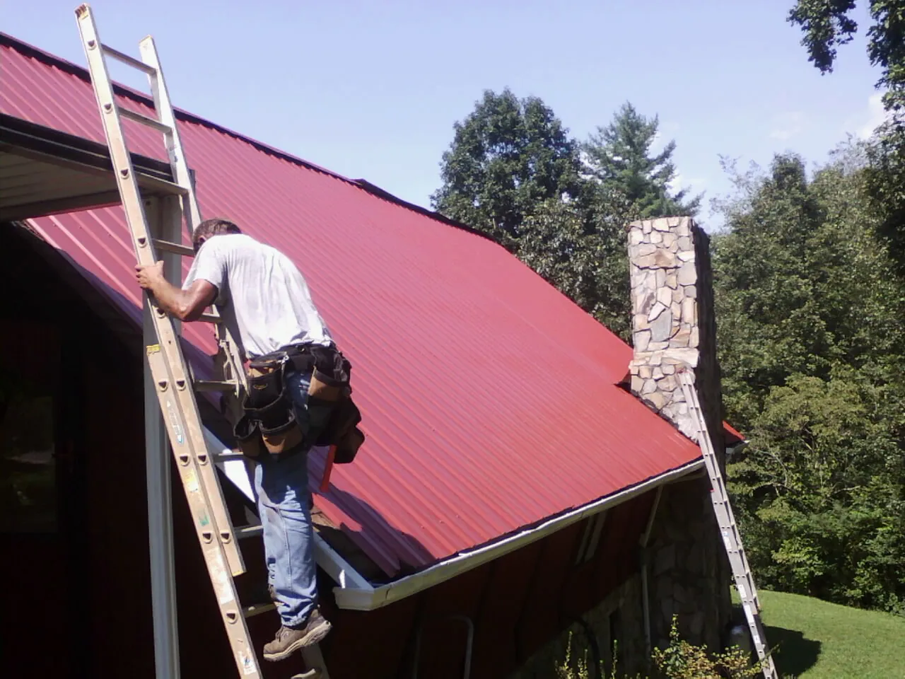 Roofer on a ladder beside a red metal roof and stone chimney — Soffit & Fascia Repair by Roofing Brothers in Klickitat